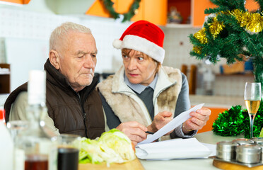 Upset elderly man with his wife reading documents at home table decorated for Christmas