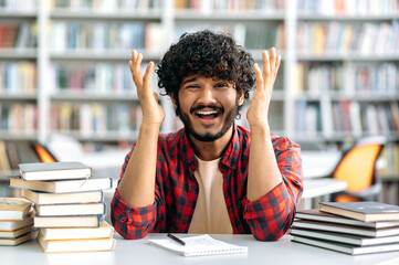 Puzzled confused indian or arabian guy, a university student, sit at a desk in library between...