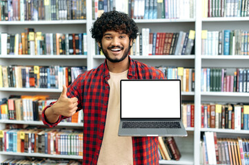 Excited millennial arabian or indian male student, shows a laptop with blank white mock-up screen, stands in a library against the background of bookshelves, looks at camera, smiles, thumb up gesture