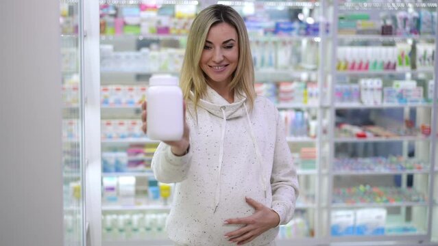 Portrait of happy pregnant beautiful woman advertising pills standing in pharmacy indoors. Front view of smiling Caucasian expectant looking at camera stretching bottle with medication