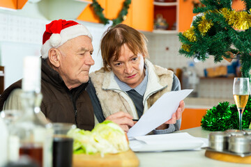 Worried mature couple sitting at table with financial documents in kitchen before christmas dinner