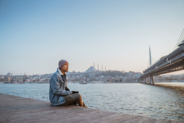 man relaxing on the side of bosphorus during sunset. asian young male with istanbul city scape at...