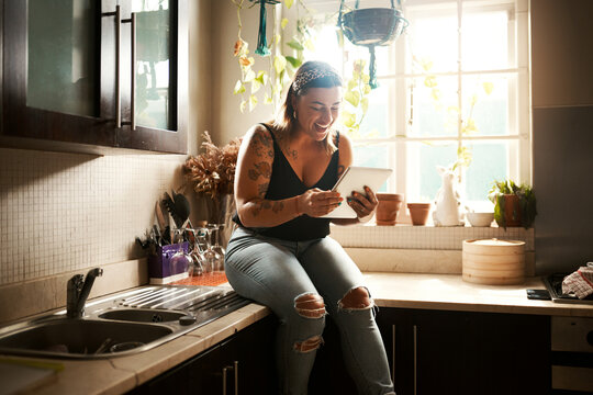 Browsing social media on tablet, searching online and reading funny stories, gossip and articles on technology app in home kitchen. Smiling and happy woman sitting on countertop and playing games