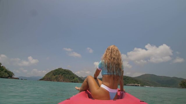 Pretty young blonde woman in stylish swimsuit and sunglasses sits on pink canoe sailing along sea against hills. Traveling to tropical countries. Attractive sexy girl is sailing on kayak in ocean.