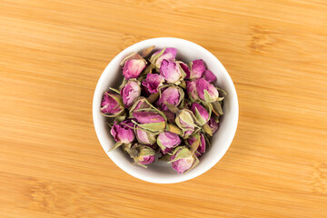 Dehydrated pink rose buds in a white bowl top down view on a wood cutting board