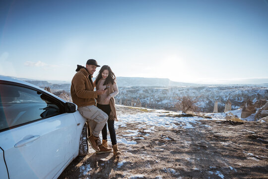 Asian Couple Doing Adventure By Car Looking At Mobile Phone With Beautiful Cappadocia Cover With Snow In The Background