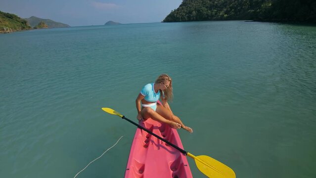Pretty sportive blonde lady in stylish swimsuit sails on pink plastic canoe along azure ocean bay at exotic resort. Traveling to tropical countries. Girl is sailing on kayak in ocean, view from above.