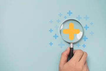 Fotobehang Hand holding magnifier glass with yellow plus sign symbol inside for focus healthcare insurance and offer positive thinking mindset of personal development concept. © Dilok