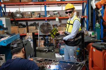 Engineer wearing helmet and hi visible vest working in factory hold laptop computer specification looking at welder prefabricated modify automatic welding support for robot arm
