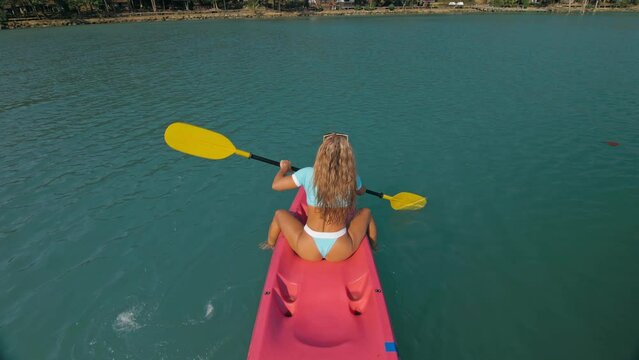 Seductive sexy blonde woman in blue swimsuit sails on pink plastic kayak along azure sea at exotic resort backside upper above view. Traveling to tropical countries. Girl is sailing on kayak in ocean.