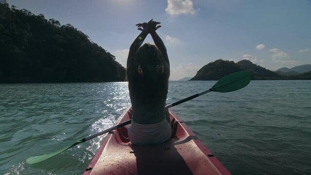 Silhouette of woman tourist raising hands while sailing in canoe along sea bay water to distant hilly island backside view. Traveling to tropical countries. Sports girl is sailing on kayak in ocean.