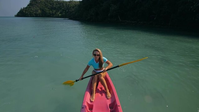 Pretty sportive blonde lady in stylish swimsuit sails on pink plastic canoe along azure ocean bay at exotic resort. Traveling to tropical countries. Girl is sailing on kayak in ocean, view from above.