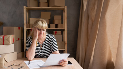 Sad depressed older woman owner of small business holding documents, looks through sales dynamics...