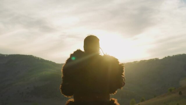 Performer raises up and taps shaman drum against mountains at sunrise backside view. Sacred spiritual ritual of Yakut folks. Highland scene