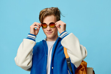 a handsome, stylish guy student with a backpack on his back in a blue bomber jacket puts on his...