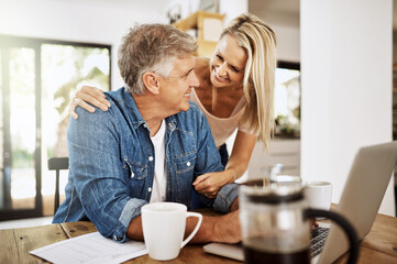 Fototapeta na wymiar Mature couple talking and browsing on a laptop while spending time together. Loving and happy wife hugging and distracting her freelancing husband while working from home at the kitchen table