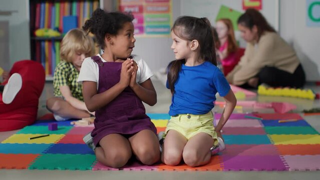 Multiracial little girls tell secrets sitting on floor in kindergarten