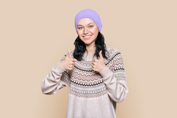 Happy young woman in winter sweater hat showing thumbs up, on brown background