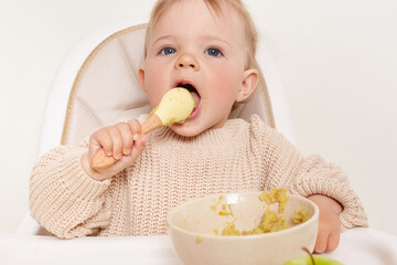 Image of funny hungry baby girl sitting in chair and eating, holding spoon in hands, keeps mouth...