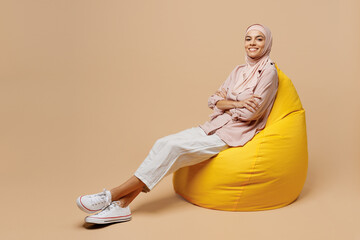 Full size side view young arabian asian muslim woman she wear abaya hijab pink clothes sit in bag chair hold hands crossed folded isolated on plain beige background People uae islam religious concept