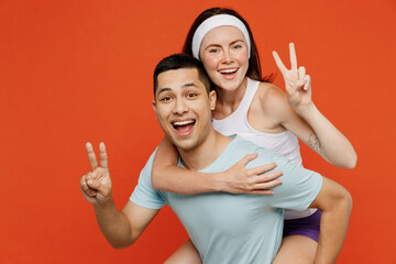 Young fitness trainer instructor sporty two man woman in headband t-shirt give piggyback ride to joyful spend show v-sign weekend in home gym isolated on plain orange background. Workout sport concept