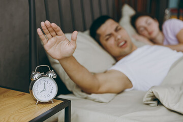 Young sad tired couple two family man woman she he wearing t-shirt pajama turns off alarm lying in...