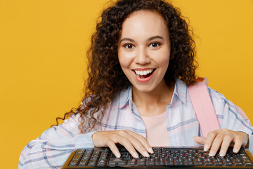 Young excited black teen girl student she wear casual clothes backpack bag typing on laptop pc keyboard look camera isolated on plain yellow color background. High school university college concept.