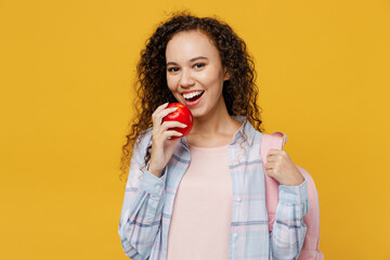 Young smiling happy cool black teen girl student she wearing casual clothes backpack bag holding eat biting red apple isolated on plain yellow color background. High school university college concept.