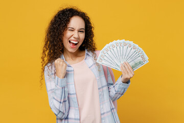 Fototapeta Young black teen girl student she wear casual clothes backpack bag hold fan cash money in dollar banknotes do winner gesture isolated on plain yellow background. High school university college concept obraz