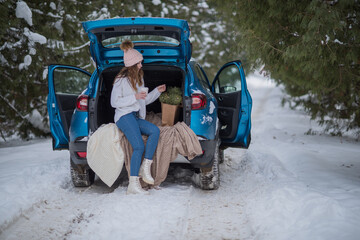 A woman with hot coffee in her hands sits in a blue car on a snowy winter day in the forest. Girl with a cup of hot coffee sits in a car with an open trunk