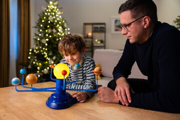 Fototapeta premium Caring dad and curious ambitious son spend time together by Christmas tree, relax playing with solar system toy, men look at rotating planets