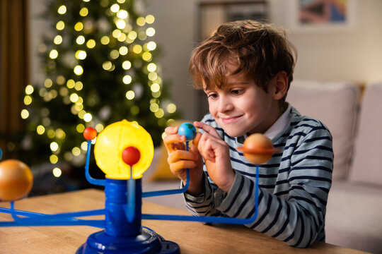 A child plays by a Christmas tree lit with lights, a school-age boy looks at a toy planetary system he built himself, fun combined with learning, the kid holds the planet