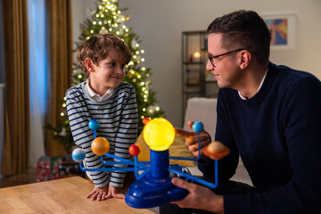 Son is spending time with dad men are sitting on couch playing with a model of planetary system guy is explaining to boy about construction of cosmos using toy as example he is naming planets