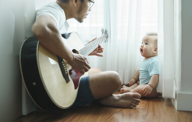 Asian Father sitting singing and playing acoustic guitar with little Cute baby infant boy together...