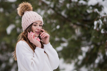 portrait of a happy young girl with a hat and a white sweater in winter in a forest or park, holding pink macaroons in her hand, a happy girl walks in nature in winter, winter