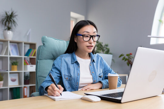 Distance Learning. Young beautiful Asian female student taking an online exam. Sitting in glasses and jeans at the table at the computer, making notes in a notebook.