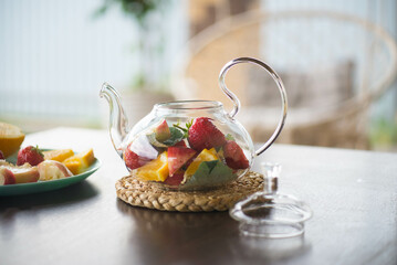 A glass teapot with fruits and berries, mint leaves on the table outside. Summer fruit tea.
