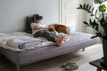 Single middle-aged woman spends weekend time playing VR games wearing VR glasses lying on bed. Lonely relaxed female in nightwear has playing games addiction staying home Dog only friend sleeps nearby