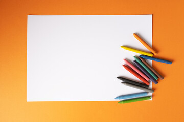 A blank white sheet of paper with wax crayons lies on a orange background