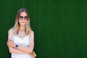 Lifestyle Portrait of stylish young caucasian woman in white casual clothes and sunglasses looking camera. Person standing near green wall background. Mock up with copy space