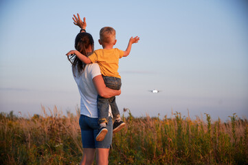 Back view of woman with kid waving hands to flying commercial airplane in the sky at sunset....
