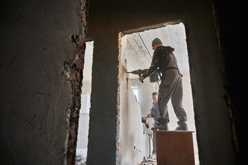 Bottom view of two builders deconstructing interior partition. One worker standing on desk with...
