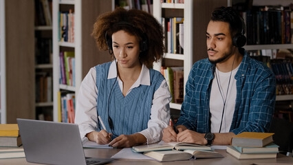 Two students sit at desk in university library listen to teacher in headphones on laptop study write down information in notebook lecture distance learning online via video conference webinar