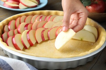 Woman putting apple slices into dish with raw dough at wooden table, closeup. Baking pie