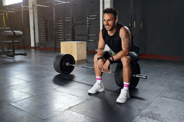 Fototapeta na wymiar Little break. Handsome Young Man in Sportswear Looking Away with Smile and Relaxing while Sitting at Gym at the Dumbbell. Sport and Recreation Concept 