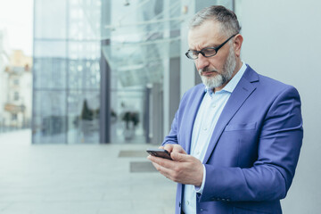 Obraz na płótnie Canvas Serious and pensive senior gray-haired man outside office building looking at smartphone screen reading news, businessman in glasses and business suit thinking about decision holding phone in hands