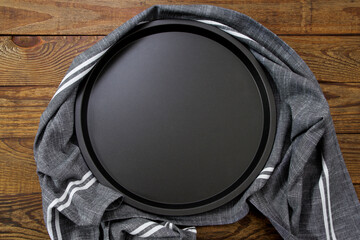 Baking pizza tray and grey tablecloth on brown wooden background. Empty black oven plate for...