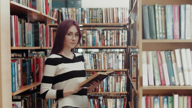 Young free Caucasian woman in glasses with purple hair stands in the library with a book and looks at the camera. Self-education library