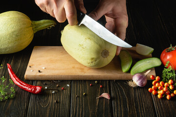 Fototapeta Hands of a cook with a knife cutting vegetable marrow on a wooden cutting board before preparing vegetarian food. obraz