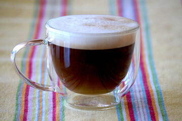 Doppelwandige Kaffeetasse aus Glas mit Kaffee geschäumter Milch steht im Vordergrund auf buntem...